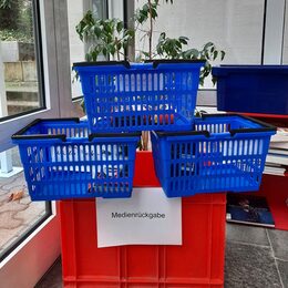 Drei blaue Ausleihe-Körbe auf einer roten Kiste zur Rückgabe der Medien